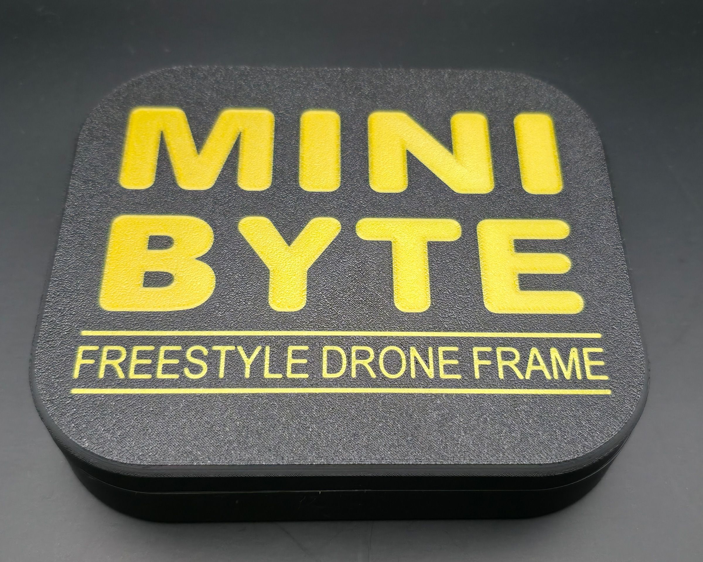 Víko 3D tištěného boxu Minibyte s vystouplým žlutým nápisem MINI BYTE a podtitulem FREESTYLE DRONE FRAME na černém podkladu.