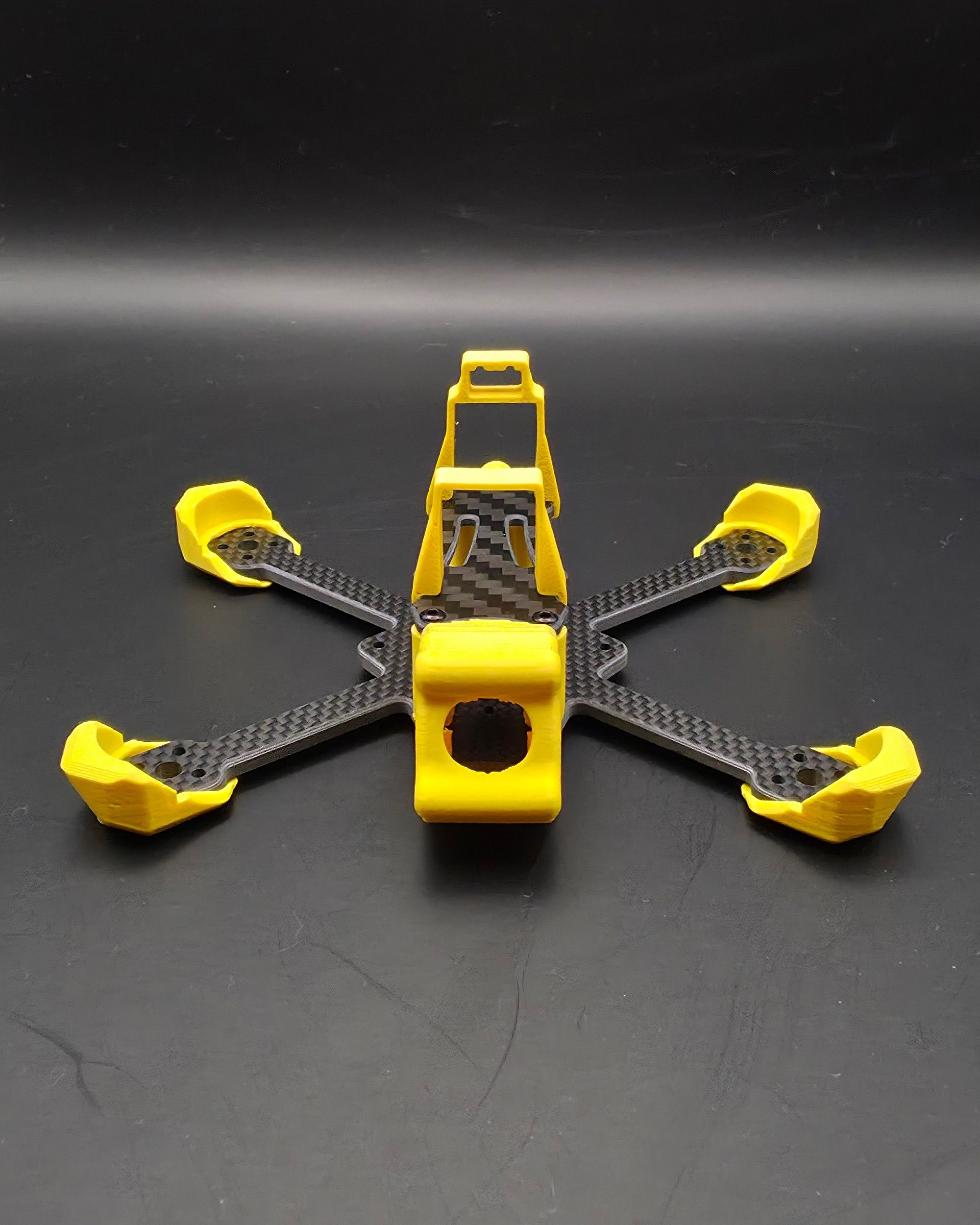 Karbonový FPV rám Minibyte 2.5 ze tříčtvrtečního pohledu — unibody karbonová deska ve tvaru X, žluté TPU chrániče ramen a žlutá TPU klec kamery s výřezem pro objektiv.